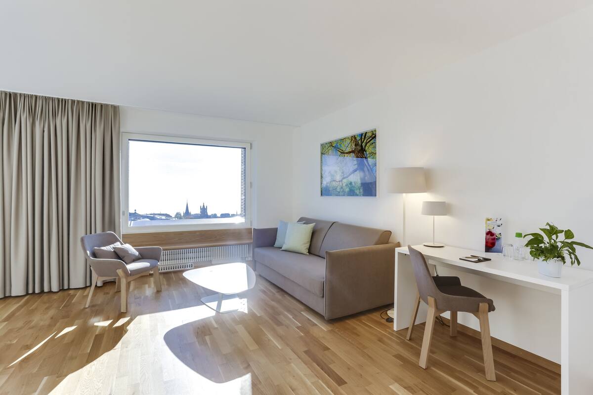 A modern living room with a sofa, two chairs, a desk, and a large window offering a city view.
