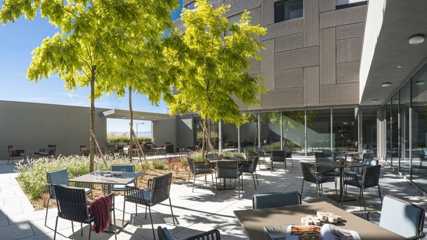 An outdoor seating area with tables and chairs, surrounded by glass walls and a building with a modern design. - Hôtel des Patients (Lausanne)