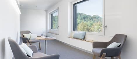 Quarto moderno com duas cadeiras cinzas, uma mesa redonda pequena e uma janela com vista para árvores.