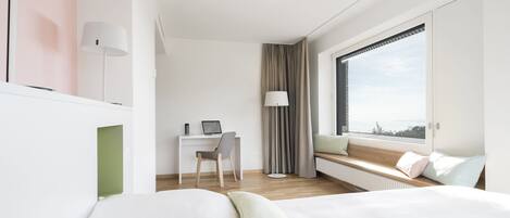Una habitación de hotel moderna con una cama, un escritorio con una computadora portátil, un asiento junto a la ventana y vistas al exterior.