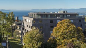 A modern building with a rooftop terrace, surrounded by trees and a view of the sea and mountains. - Hôtel des Patients (Lausanne)