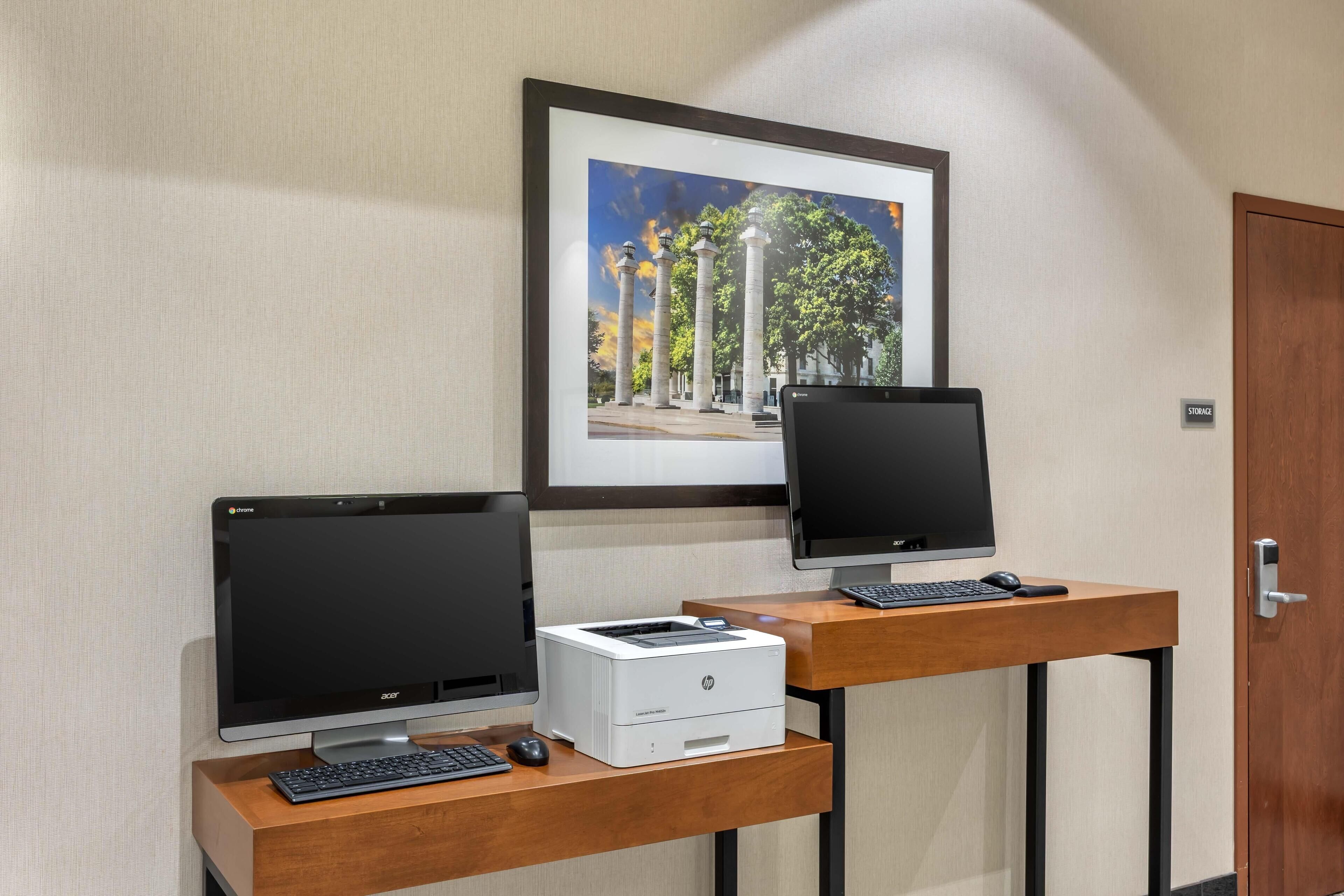 Een werkplek met twee Acer monitoren, een HP printer en een toetsenbord op een houten bureau, voor een gestructureerde wand.