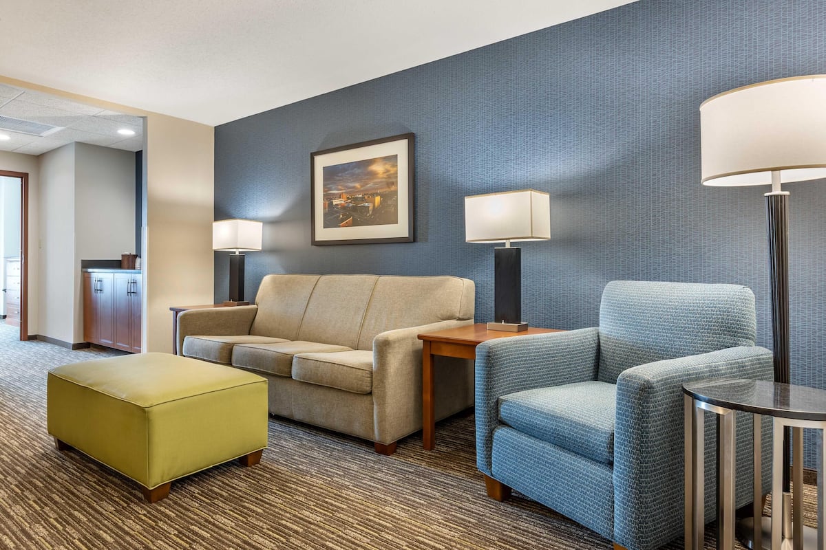 A hotel room with a beige sofa, a blue armchair, a small table, and a matching ottoman. There is a framed picture on the wall and two floor lamps.