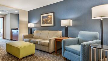 A hotel room with a beige sofa, a blue armchair, a small table, and a matching ottoman. There is a framed picture on the wall and two floor lamps.