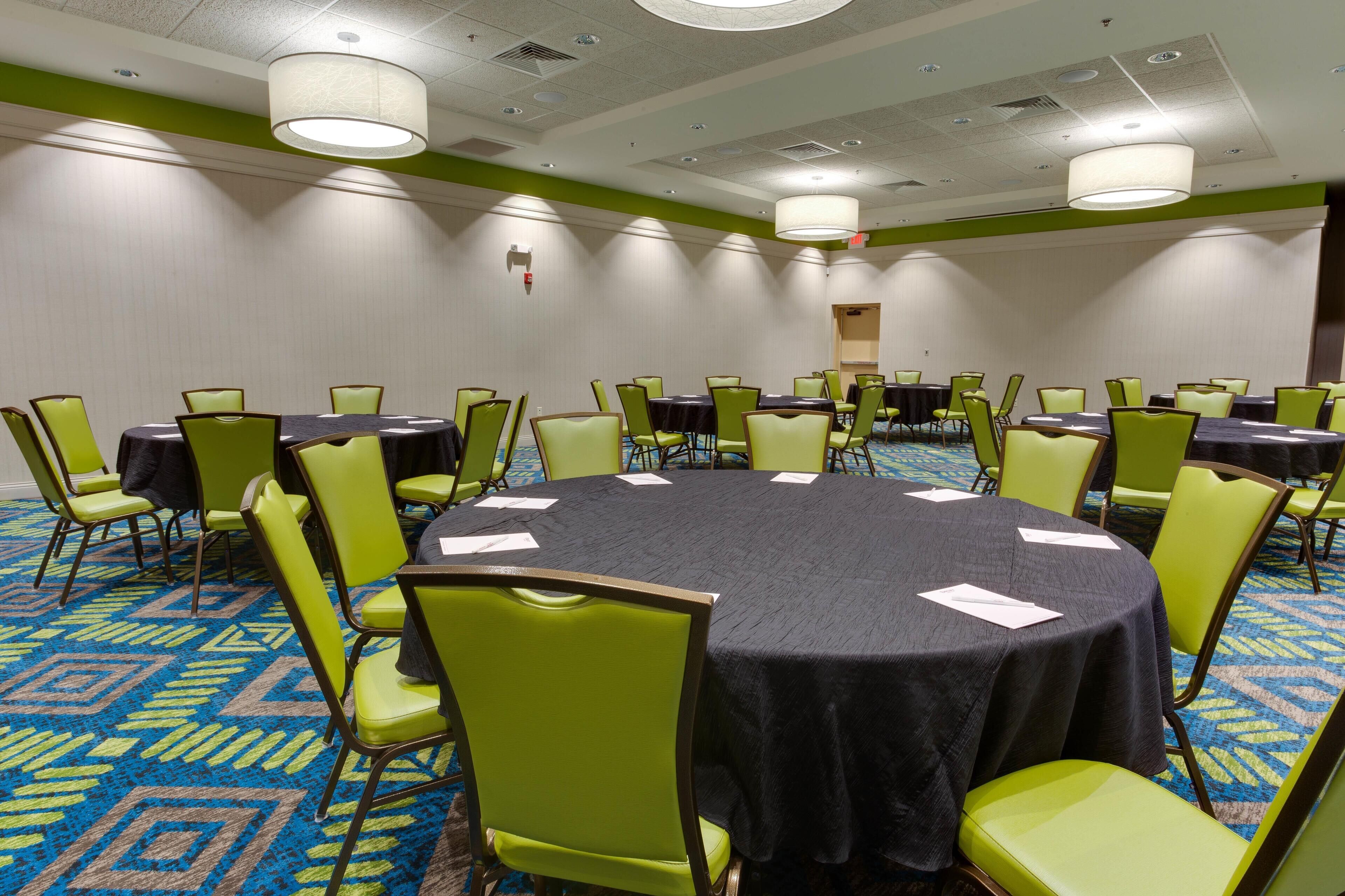 Een congreszaal met ronde tafels en groene stoelen, een vloerbedekking en verlichting in het plafond.