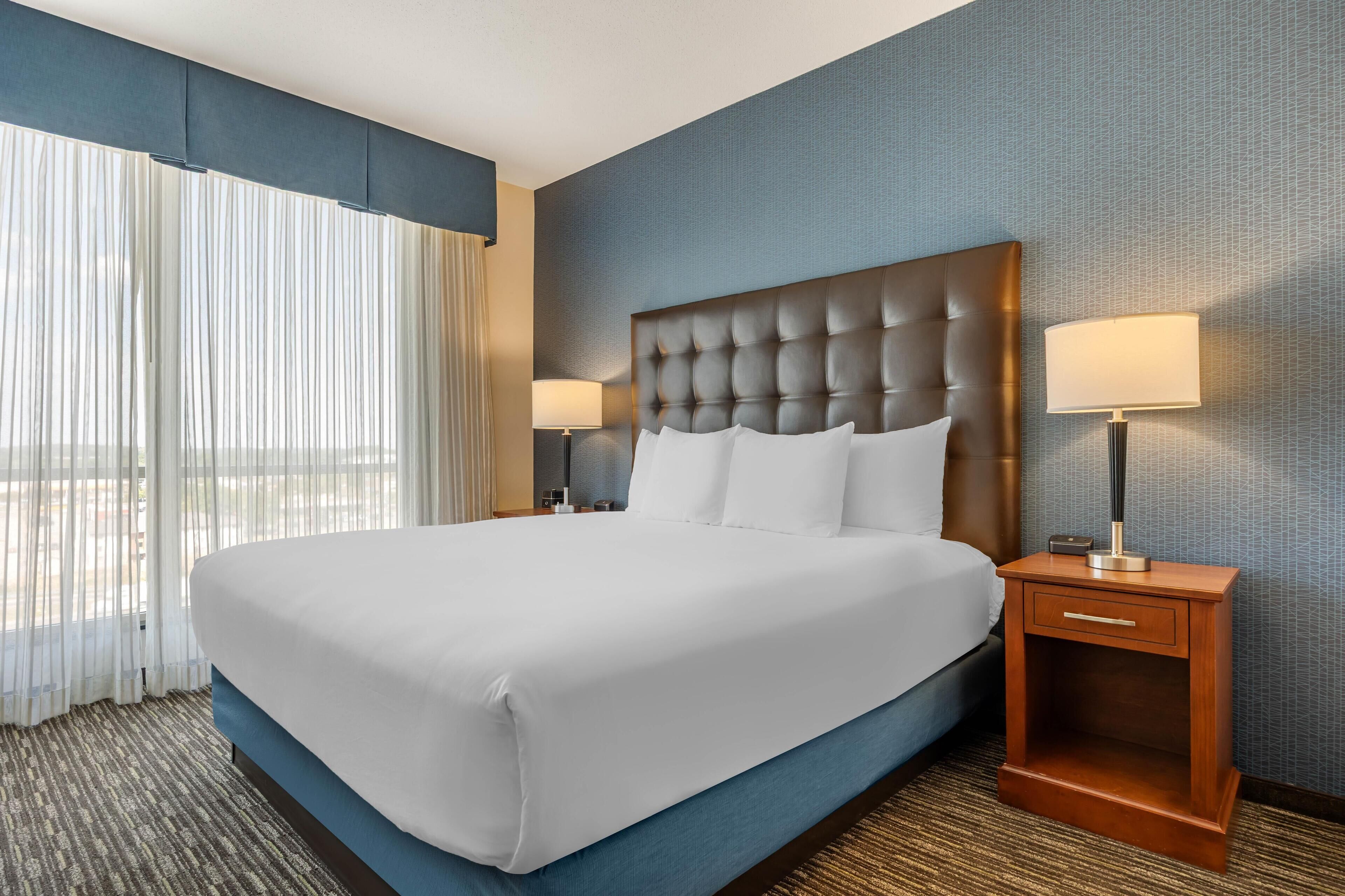A hotel room with a large bed, two bedside tables with lamps, a window with sheer curtains, and a textured blue wall.