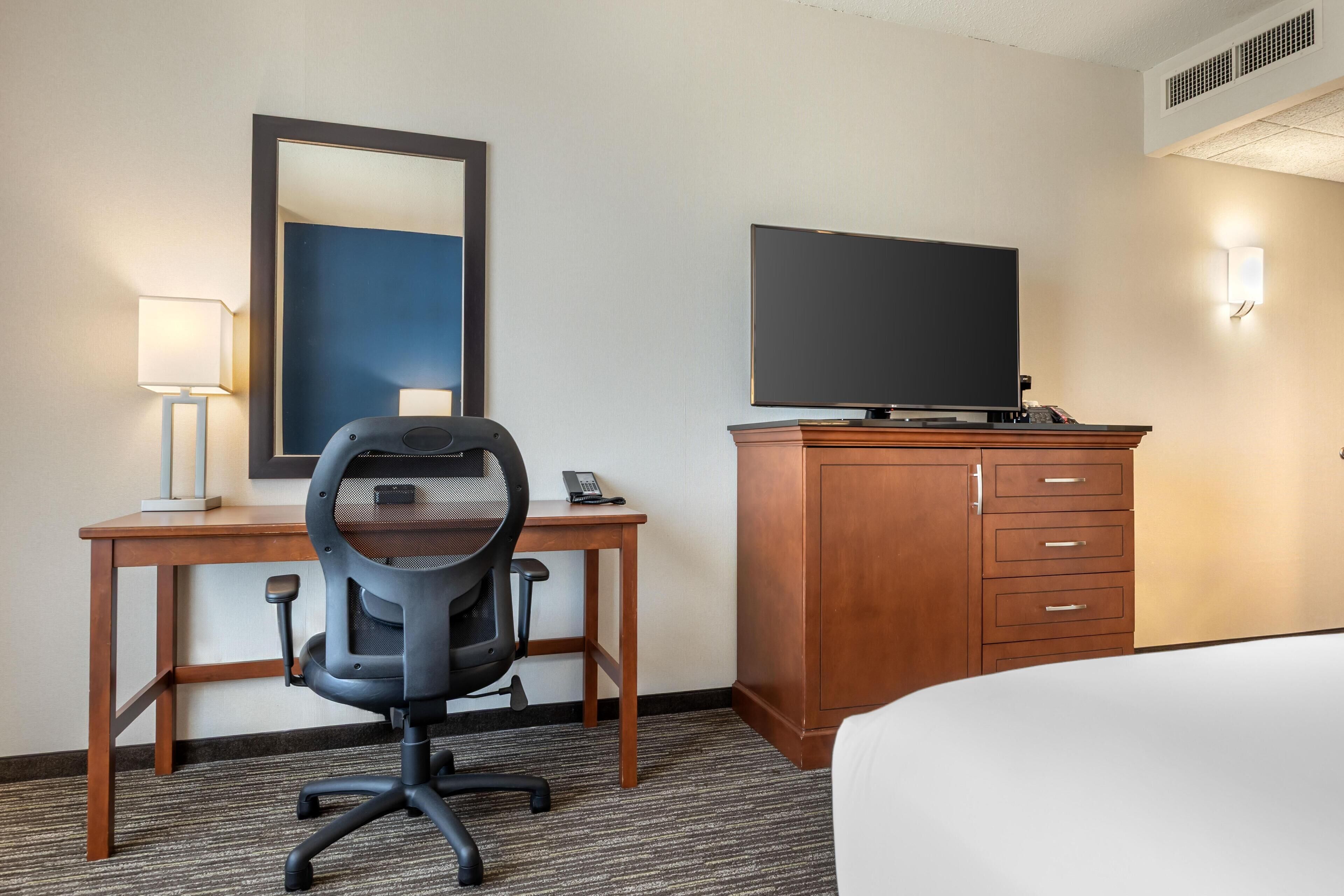 A hotel room with a bed, a desk with a chair, a television, a mirror, and a wardrobe.