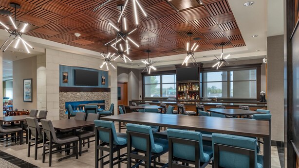 A spacious bar area with high tables and chairs, a large flat-screen TV mounted on the wall, and a bar stocked with bottles.