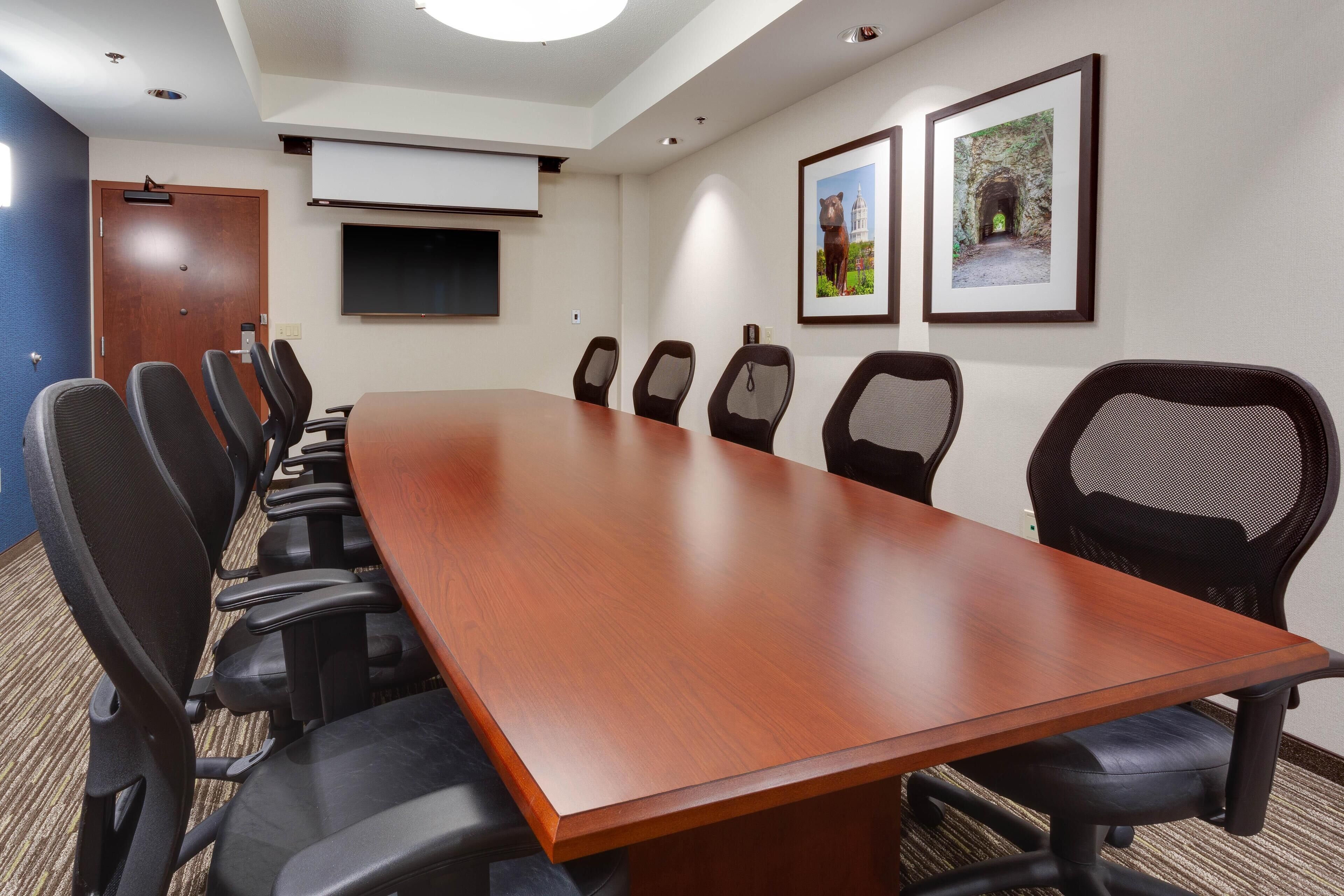 A conference room with a long wooden table, black chairs, a projector screen, and two framed pictures on the wall.