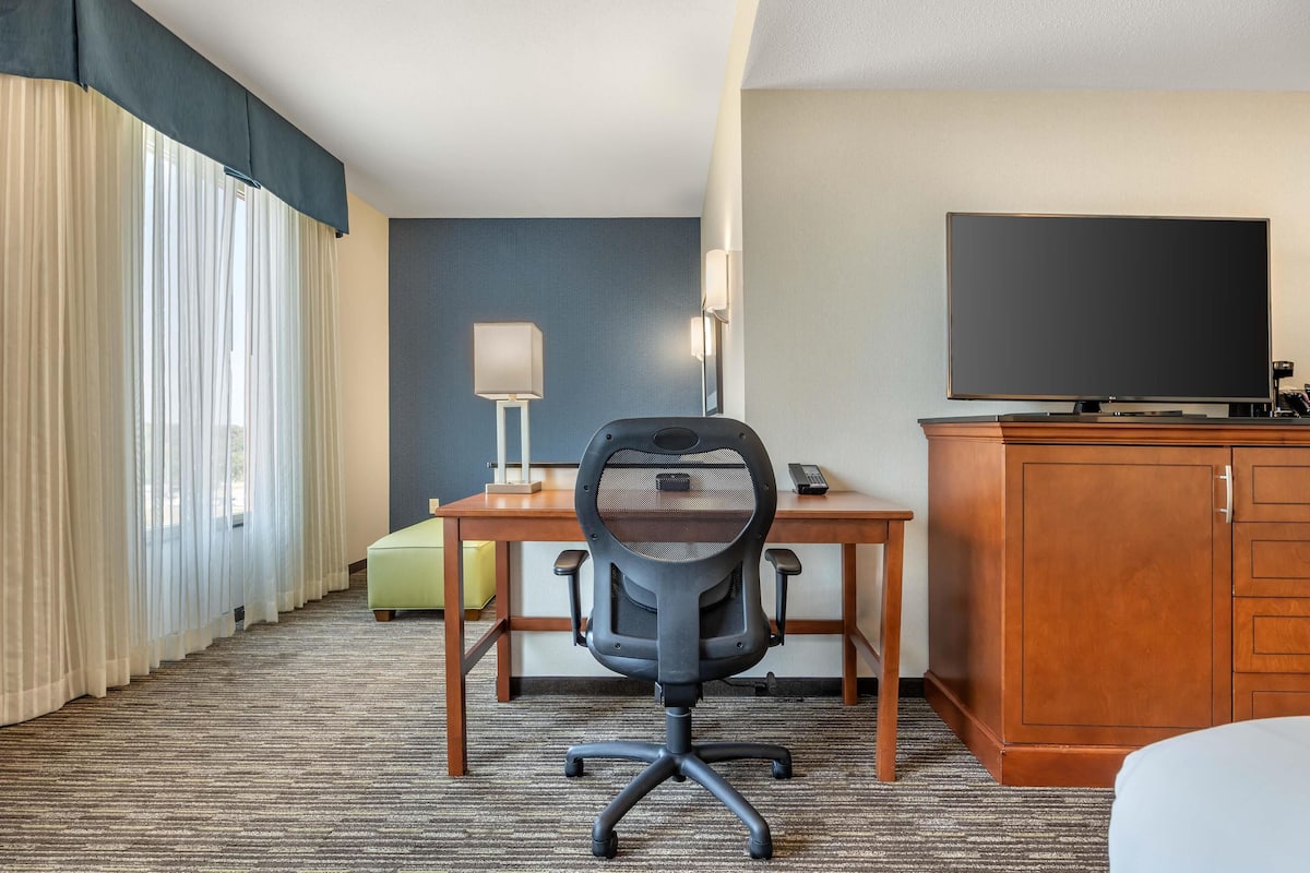A hotel room with a large flat-screen TV, a desk with a lamp, a cushioned chair, and a wooden cabinet.