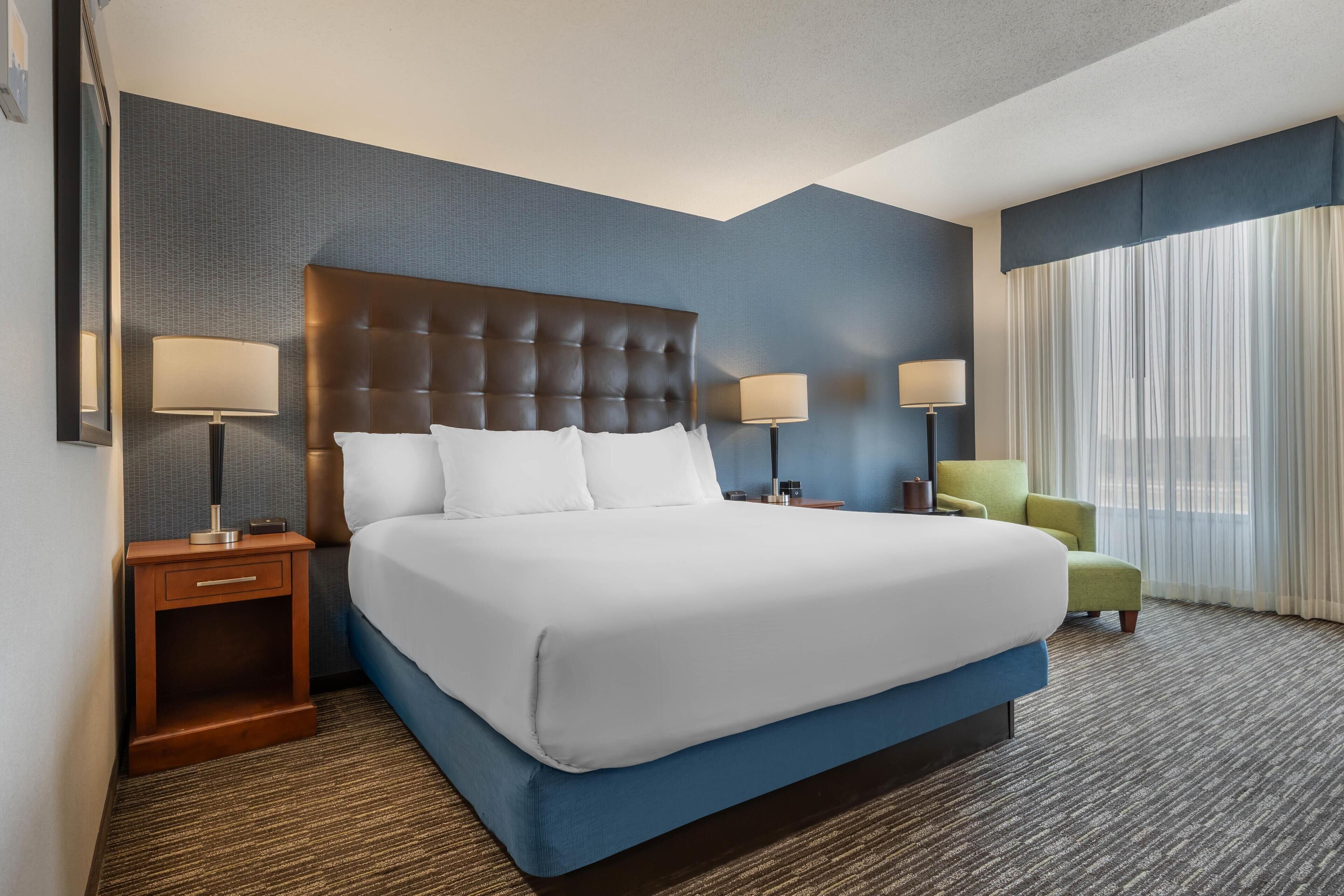 Una habitación de hotel con una cama grande, dos mesitas de noche con lámparas, un sillón verde y un ventanal con cortinas translúcidas.