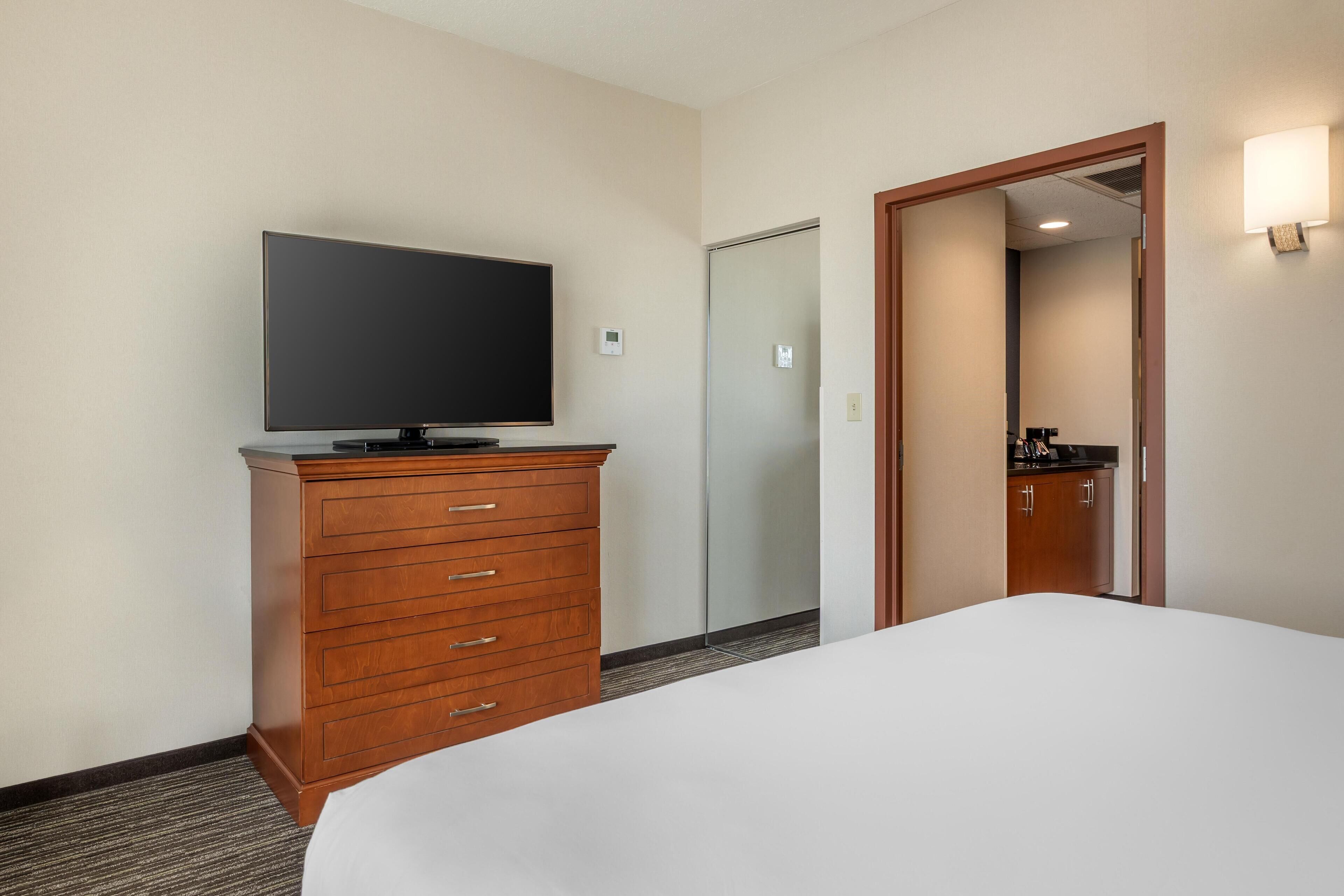 A hotel room with a bed, a wooden dresser with a flat-screen TV, a door, and a mirror reflecting a bathroom.