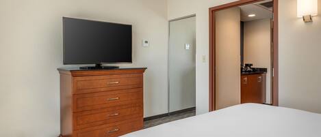 A hotel room with a bed, a wooden dresser with a flat-screen TV, a door, and a mirror reflecting a bathroom.