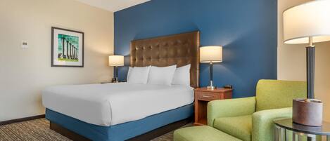 A hotel room with a large bed, two bedside lamps, a green armchair, and a small table with a lamp.