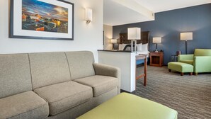 A hotel room with a sofa, a green ottoman, a desk with a lamp, and a framed cityscape picture on the wall.