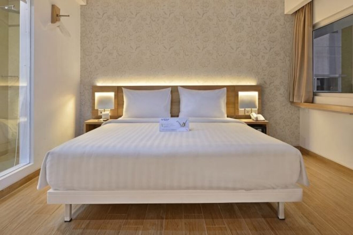 Una habitación de hotel con una cama grande, dos mesitas de noche con lámparas, un ventanal con cortinas y una pared con un diseño discreto.