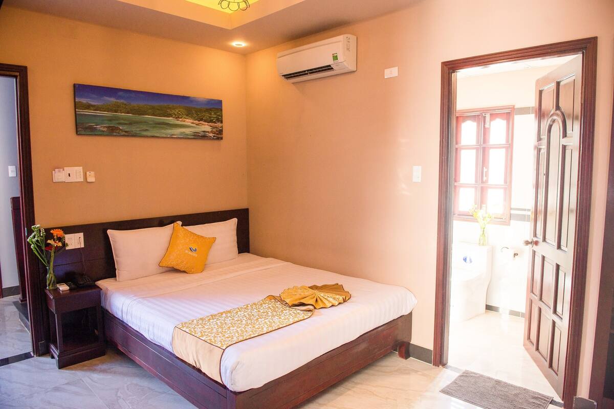 Une chambre d’hôtel avec un lit, une table de chevet, un appareil de climatisation, un tableau au mur et une porte donnant sur une autre pièce.