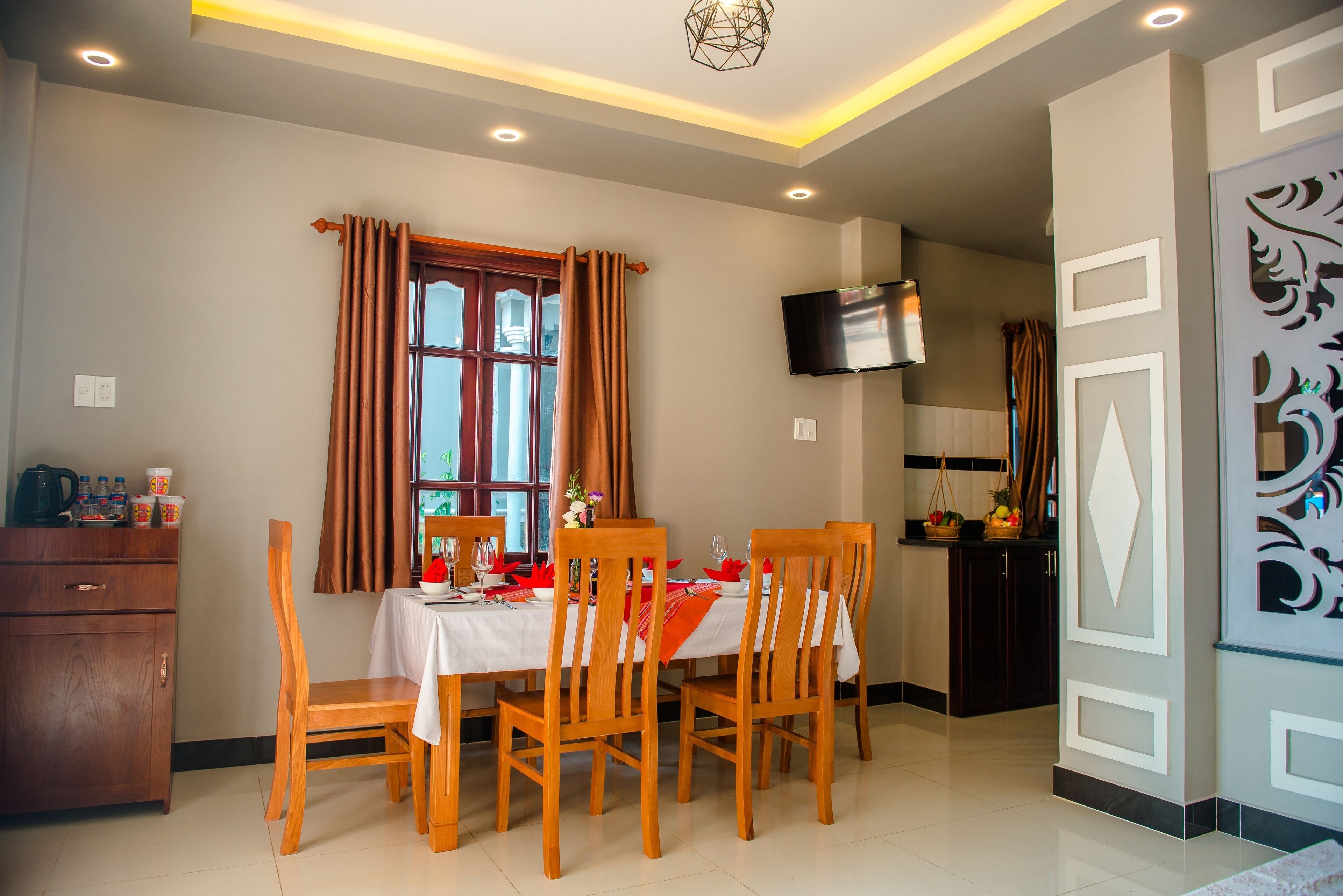 Un comedor con una mesa dispuesta para cuatro, sillas de madera, una ventana con cortinas, un televisor montado en la pared y una zona de cocina al fondo.
