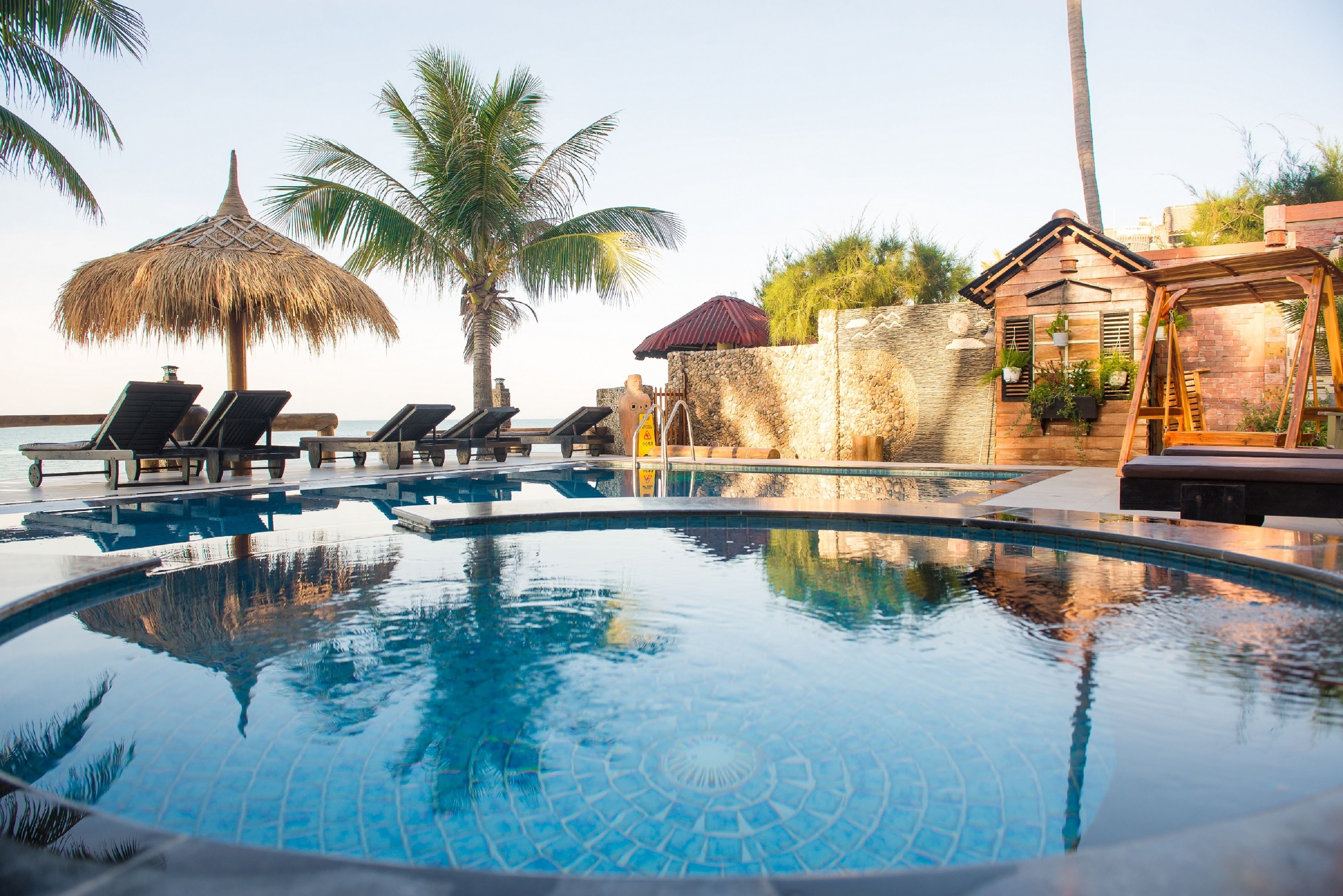 A swimming pool with lounge chairs, a thatched umbrella, and a stone wall with a wooden structure.