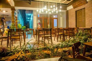 Interior de restaurante com mesas e cadeiras de madeira, luminárias esféricas pendentes e plantas em vasos.