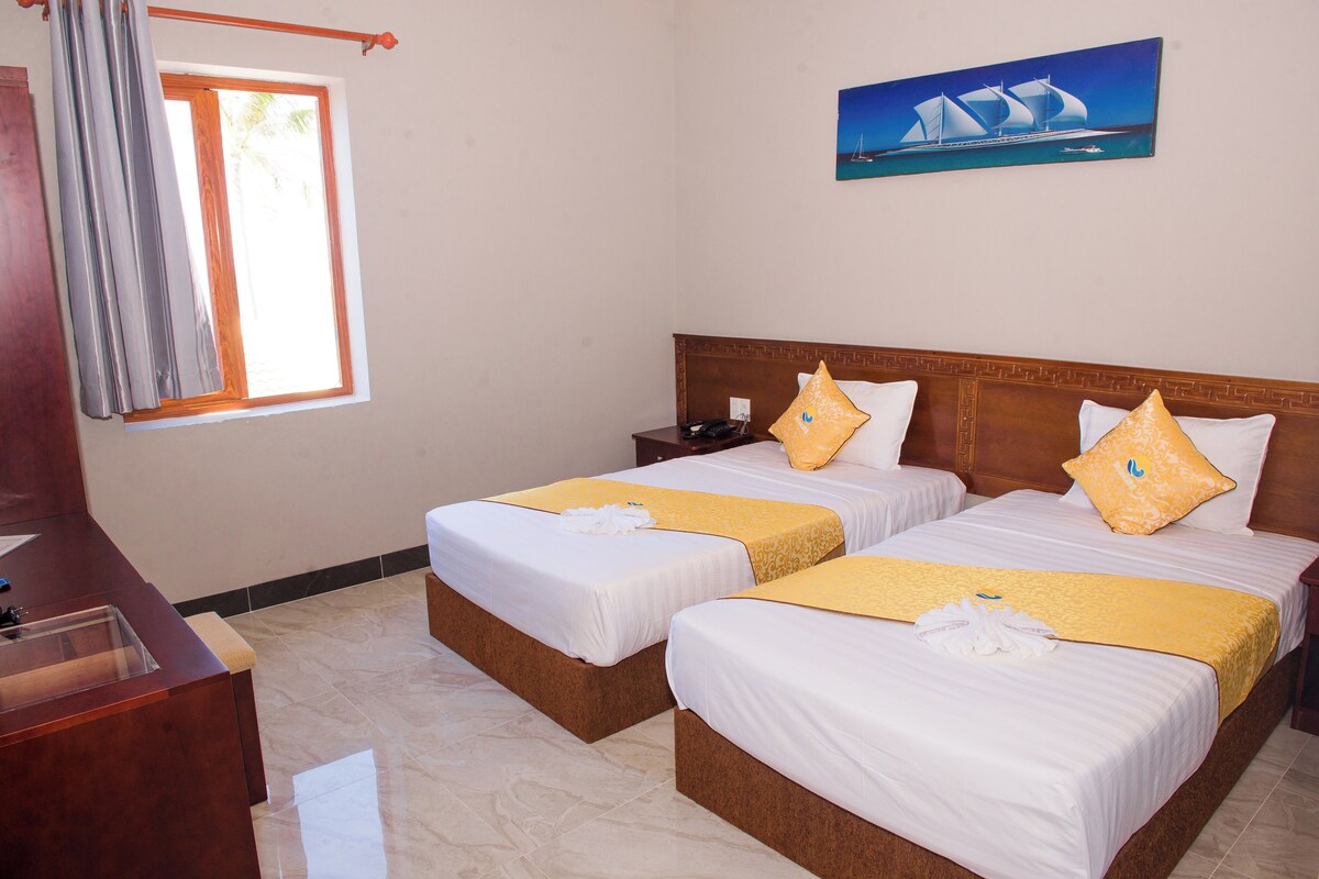 Habitación de hotel con dos camas, cabecero de madera, un cuadro de un velero, una ventana con cortinas y un escritorio con teléfono.
