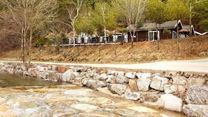 A stone-lined river with a wooden bridge, surrounded by trees and a row of cabins with sloped roofs. - Planttrees House (Pyeongchang)