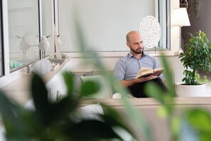 Ein Mann sitzt auf einer Couch und liest ein Buch in einem hell erleuchteten Raum mit einem großen Fenster, einer Topfpflanze und einem Wanddekor.
