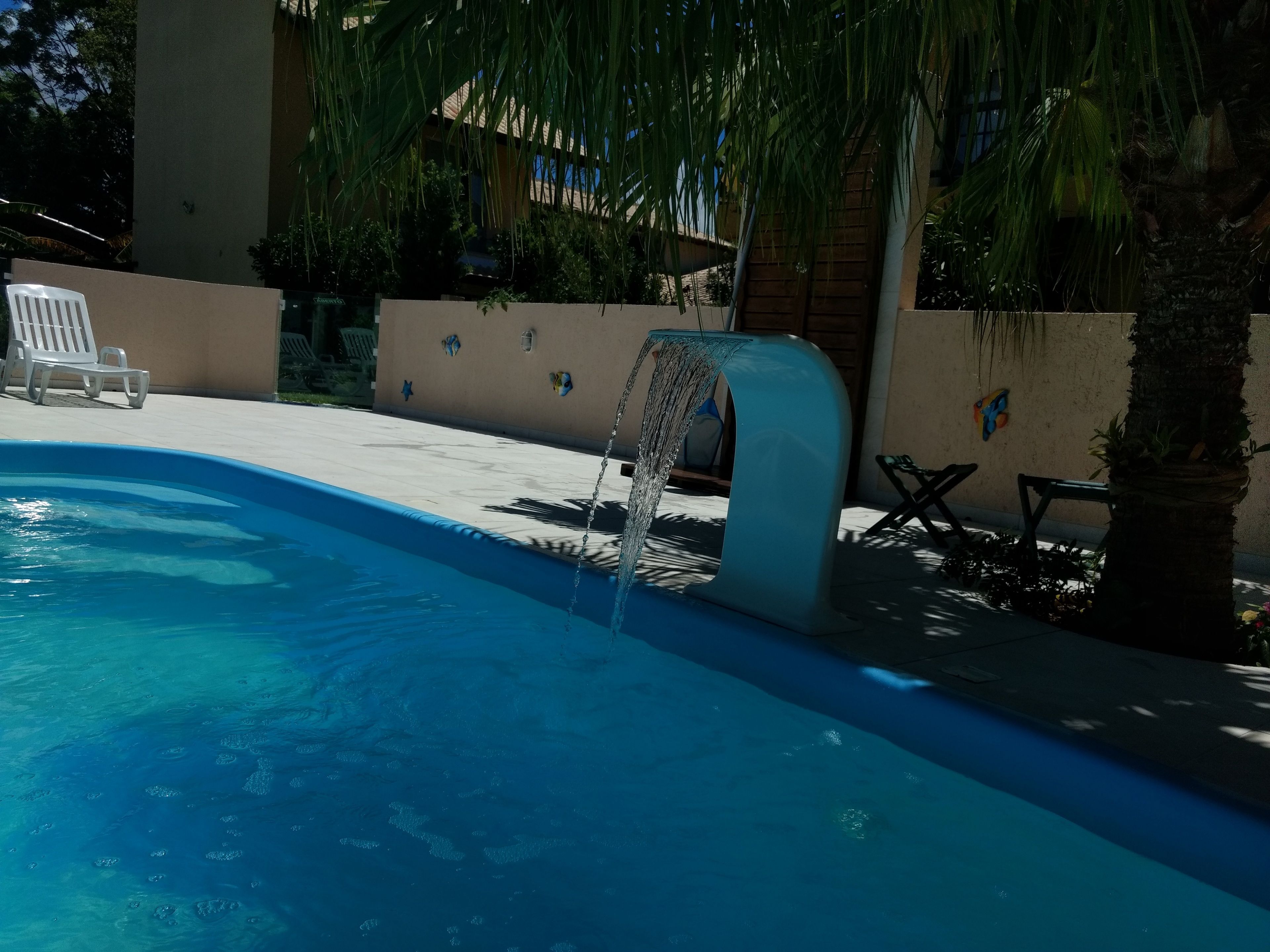 Une piscine agrémentée d’un élément décoratif aquatique, entourée d’un mur blanc orné d’éléments décoratifs, d’un palmier et d’une chaise longue.