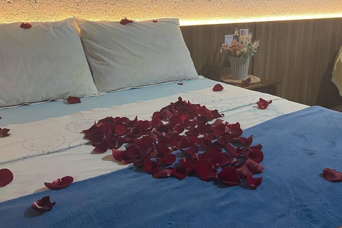 A bed with rose petals scattered on it, a window with a view of the landscape, and a small table with a vase of flowers.