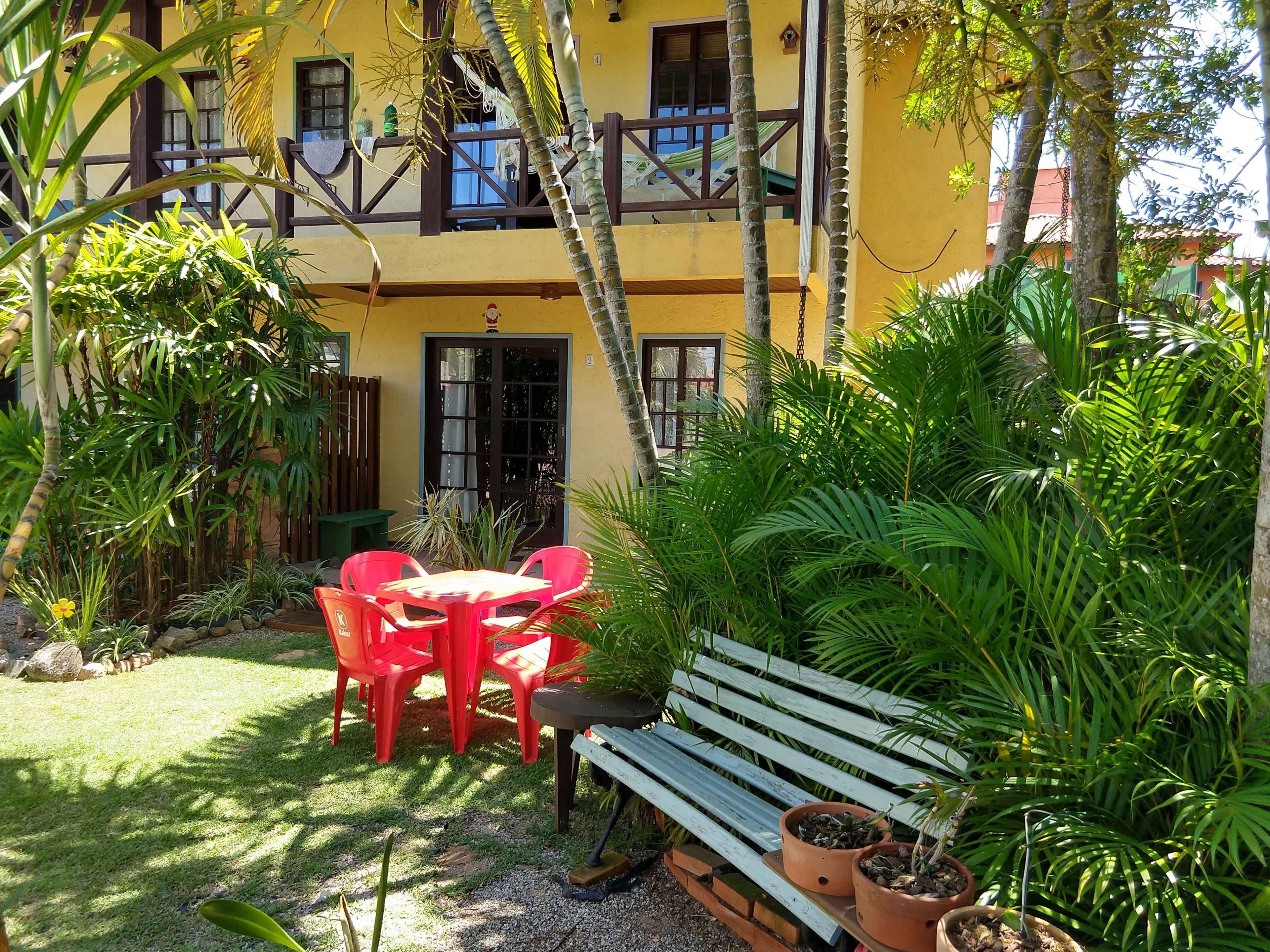 A yellow building with a balcony, surrounded by lush greenery and a small outdoor seating area with a table and chairs.