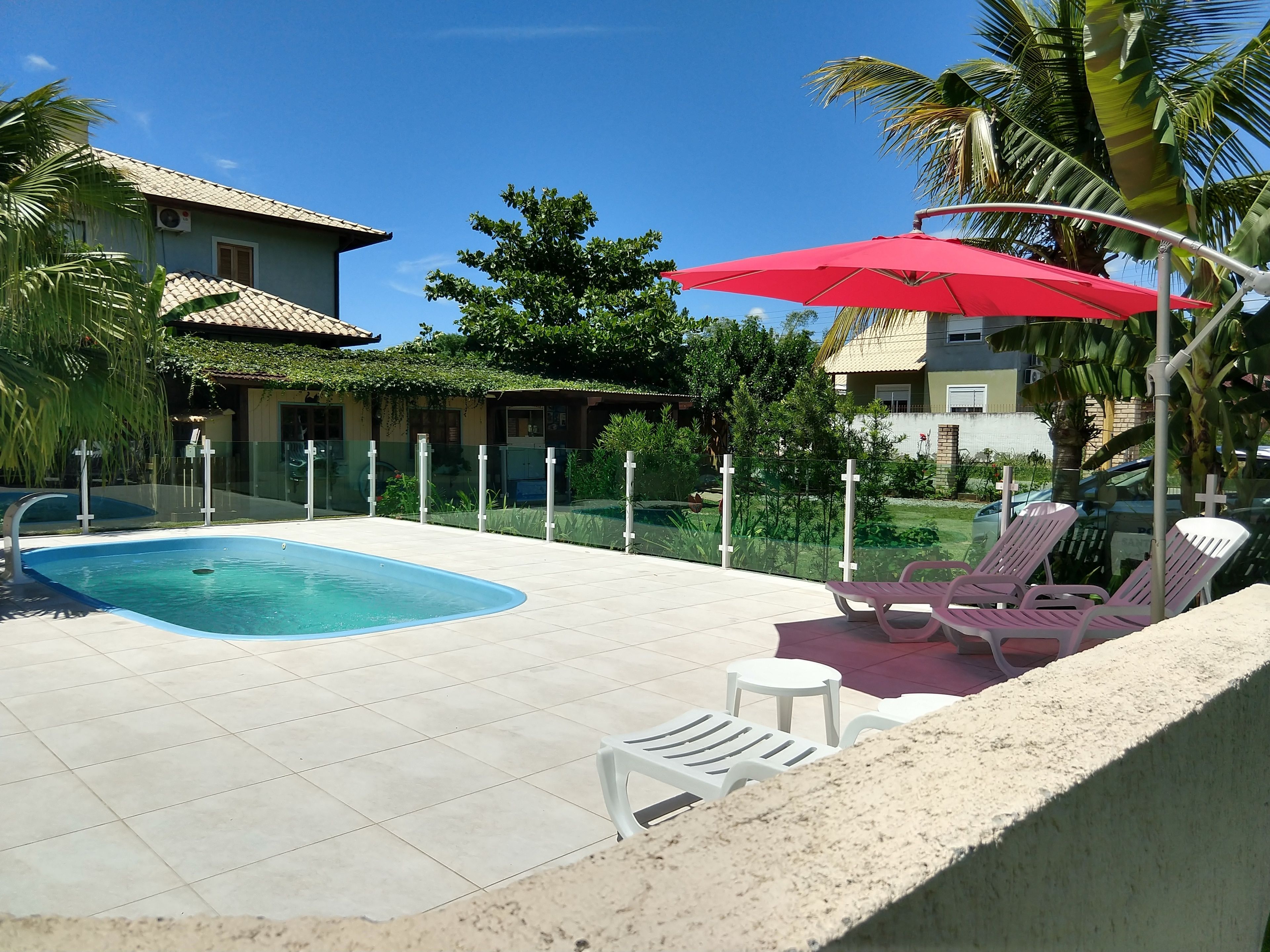 Una piscina con borde de baldosas, rodeada por una cerca baja, y una sombrilla roja que da sombra.