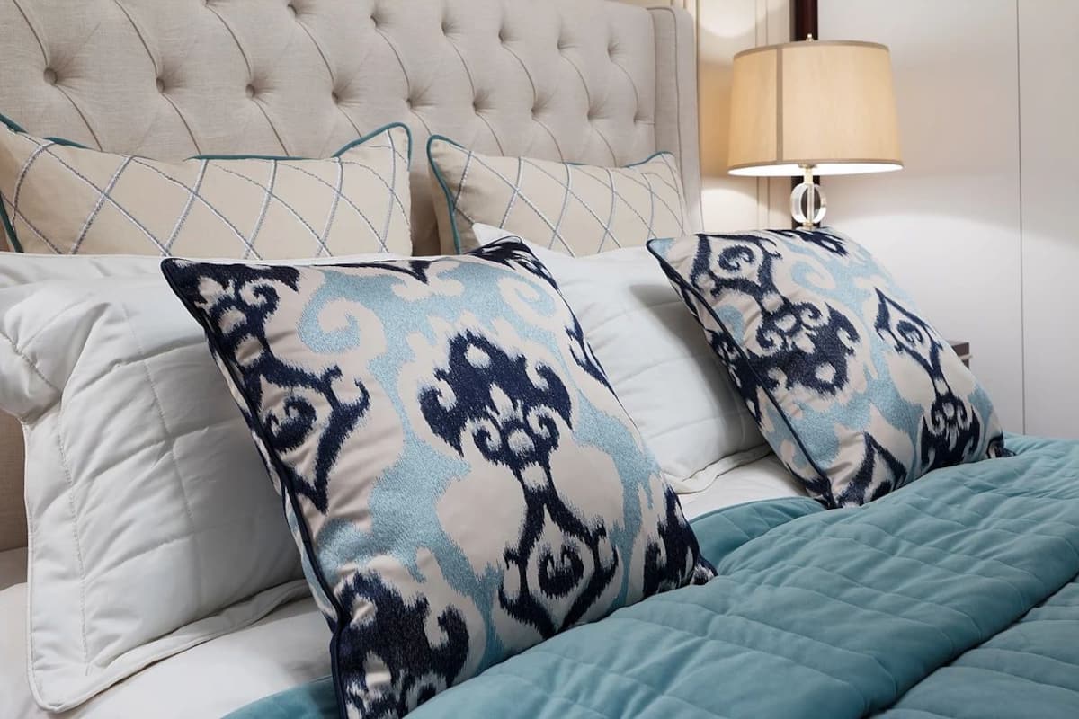 A bed with a tufted headboard, a teal quilted bedspread, and patterned pillows.