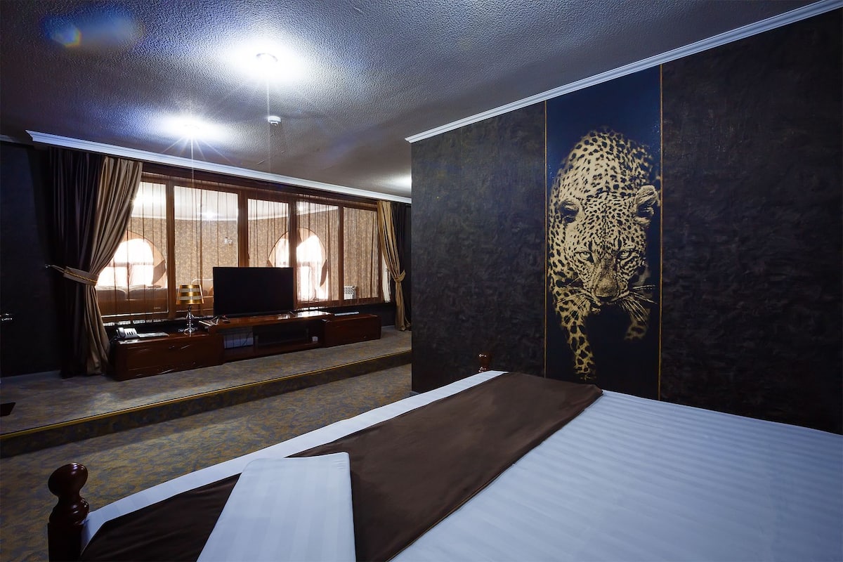 A hotel room with a bed, a TV, a large window with curtains, and a leopard print wall.