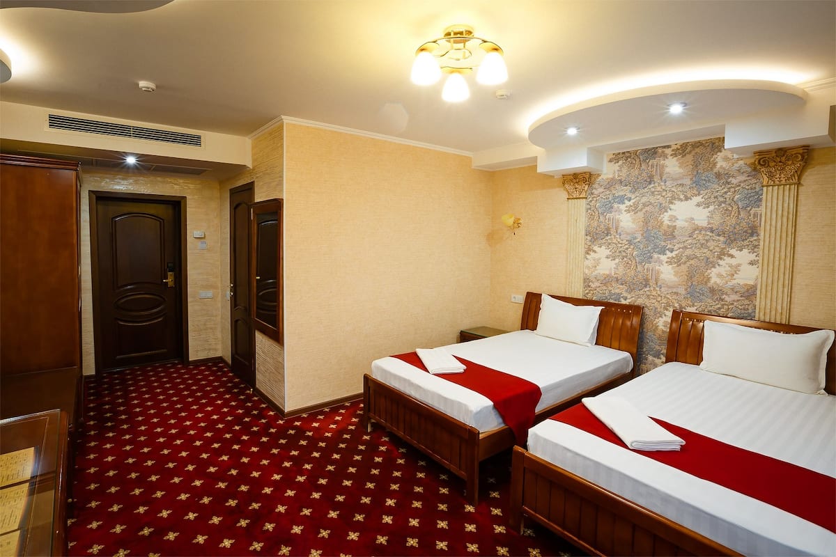 A hotel room with two beds, a red carpet, a wooden wardrobe, and a mural on the wall.