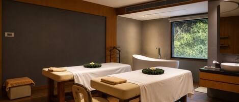 A modern spa room with two massage tables, a wooden floor, a large window with a view of greenery, and a bathroom with a sink and mirror.