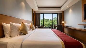 A hotel room with a large bed, a desk, a chair, a TV, and a balcony with a view of trees.