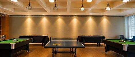 A pool table in the center with four surrounding tables, overhead lighting, and a wooden floor.