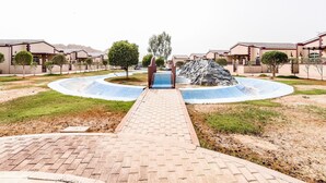 A pathway leading to a pool area surrounded by buildings with a consistent architectural style, a central tree, and a rock formation. - OYO 231 Holiday Arabian Resort (Sayh Mudayrah)
