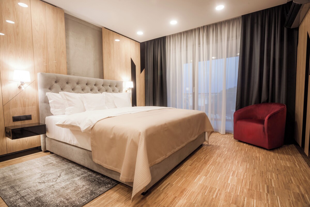 A modern hotel room with a large bed, a red chair, and a view of the city through sheer curtains.