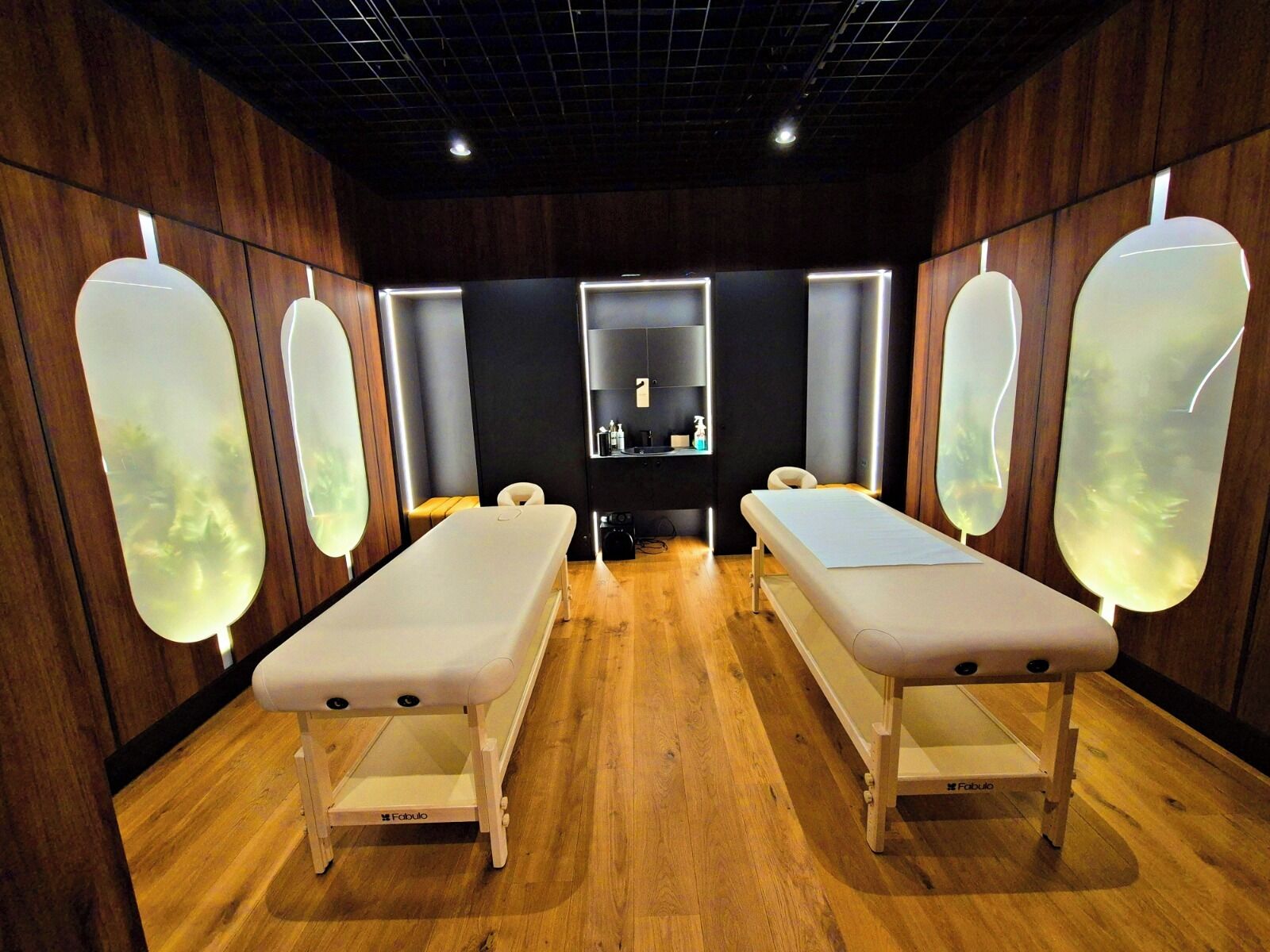 Une pièce équipée de deux tables de massage, d’un plancher en bois et d’un comptoir central agrémenté d’un miroir et d’étagères.