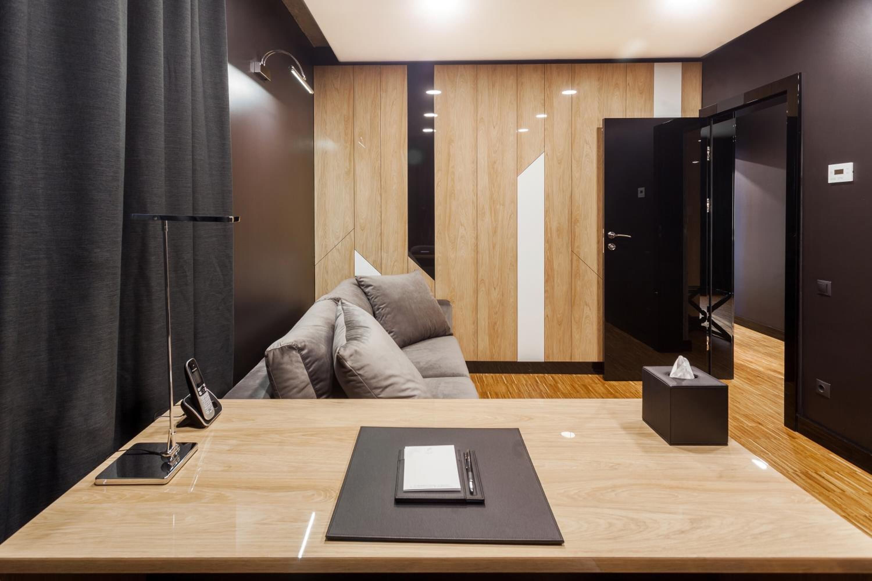 Une chambre d’hôtel moderne avec un plancher en bois, un canapé, un bureau avec un téléphone et un bloc-notes, et une porte dotée d’un miroir.