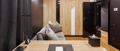 Una habitación de hotel moderna con suelo de madera, un sofá, un escritorio con teléfono y libreta, y una puerta con espejo.