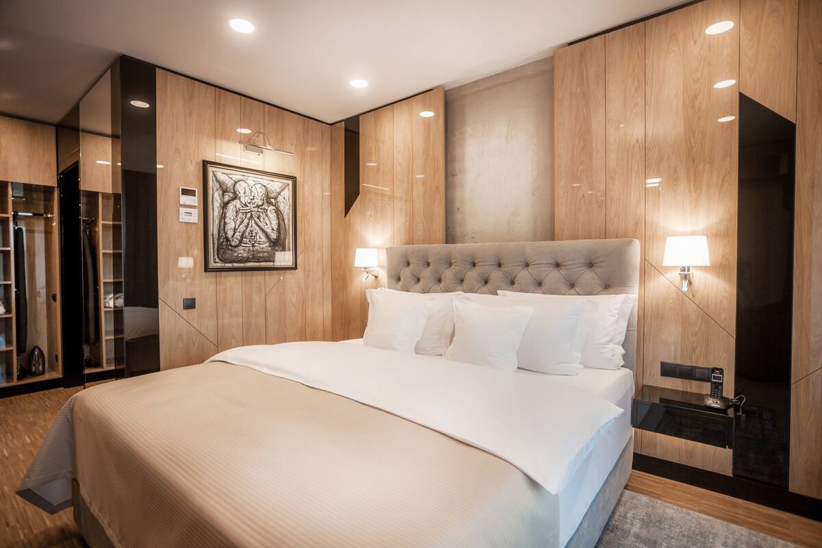 A modern bedroom with a large bed, wooden paneling, a framed artwork, and a bedside table with a lamp.