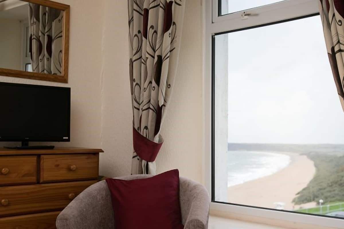 A bedroom with a bed, a TV on a wooden dresser, a chair with a red pillow, and a window with a beach view.
