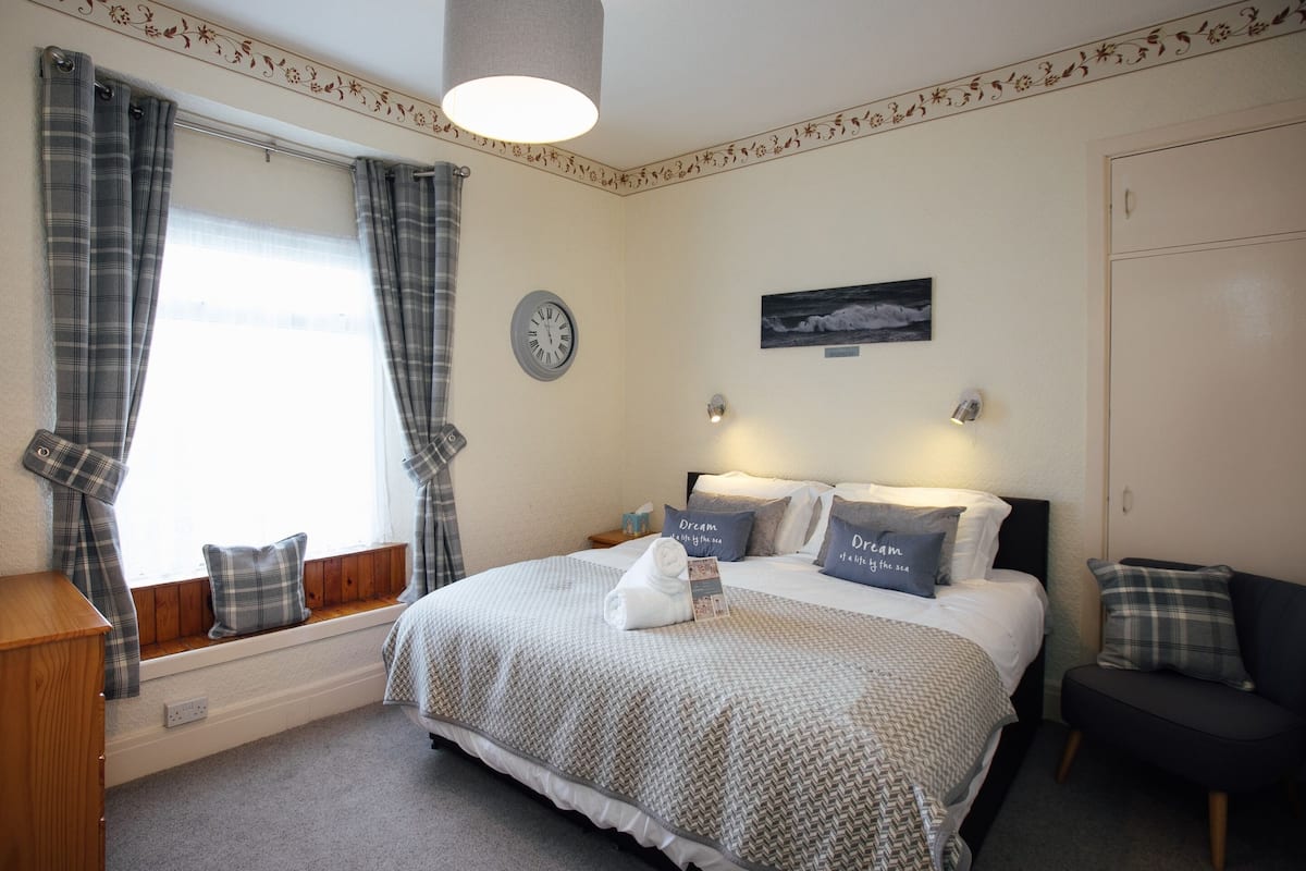 A neatly arranged bedroom with a bed, two bedside tables, a clock, and a window with curtains.