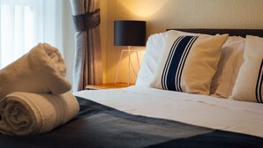 Una cama bien hecha con almohadas a rayas blancas y azul marino, una toalla enrollada y una lámpara de noche sobre una mesita de madera.