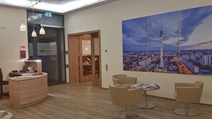 A hotel reception area with a wooden reception desk, a seating area with two chairs and a table, and a large cityscape painting on the wall. - Hotel Vita Berlin (Berlin)