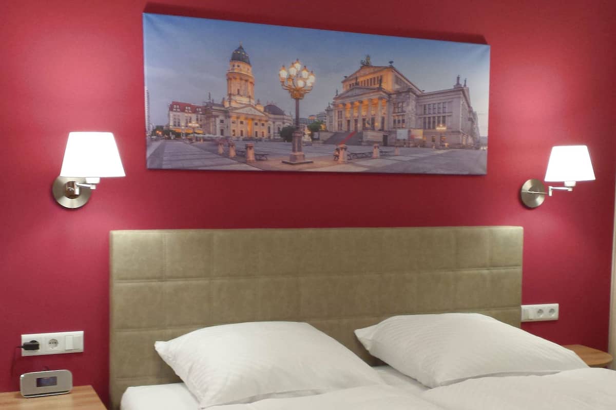 A hotel room with a bed, two bedside lamps, a painting of a cityscape, and a red wall.
