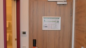 A wooden door with a control panel and a sign, an air conditioning unit above, and a red door handle. - Hotel Vita Berlin (Berlin)