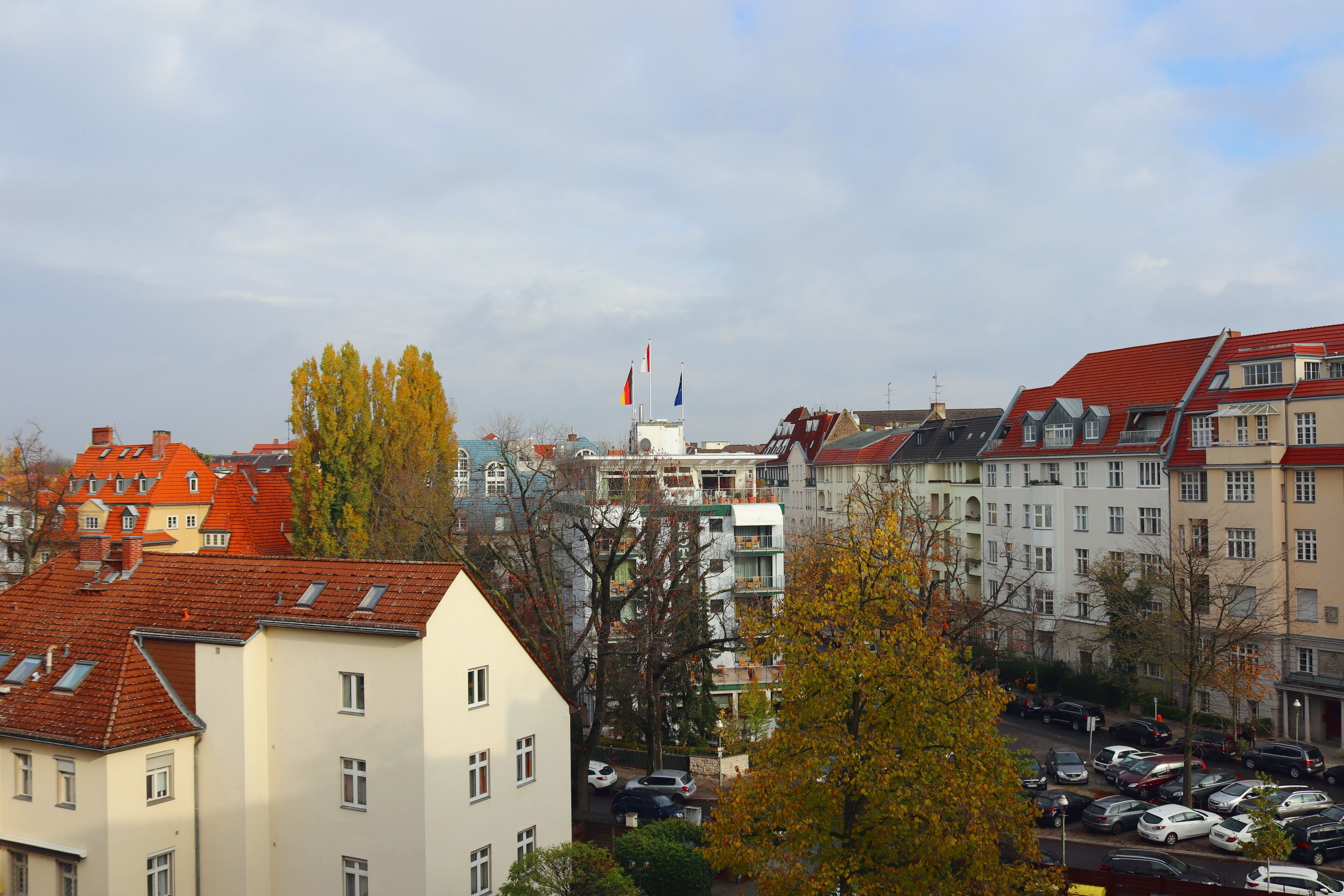 A row of multi-story buildings with red roofs and balconies, a parking lot with cars, and autumn-colored trees.